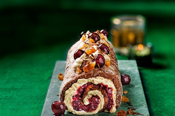 A teal board topped with a chocolate roulade. The roulade is filled with cream and whole cherries