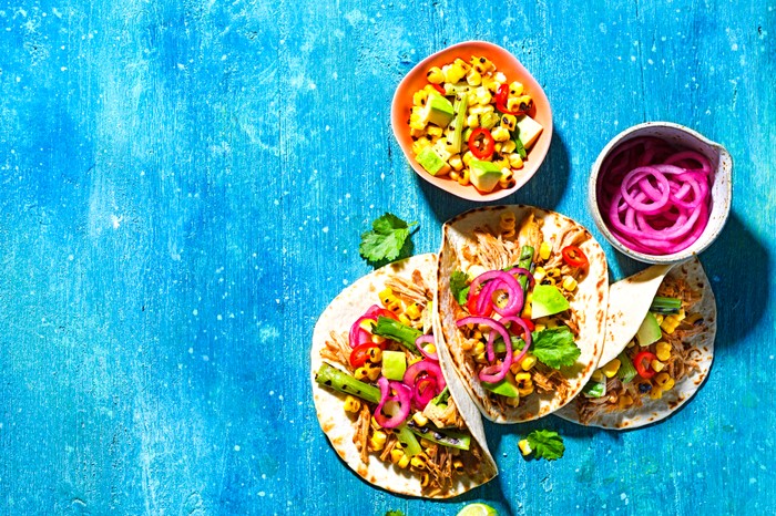 Three tacos filled with pulled pork, charred corn and pink onion on a bright blue background