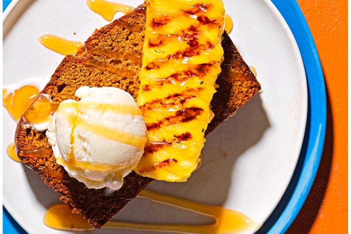 A slice of ginger and rum cake topped with a scoop of ice cream and a wedge of pineapple, on a blue plate with an orange background