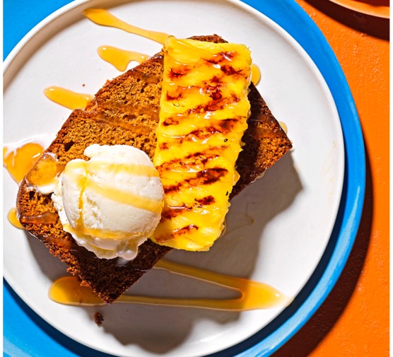 A slice of ginger and rum cake topped with a scoop of ice cream and a wedge of pineapple, on a blue plate with an orange background