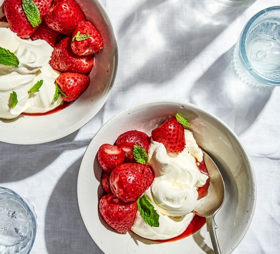 Two white bowls filled with strawberries, cream and green herbs on a white table cloth