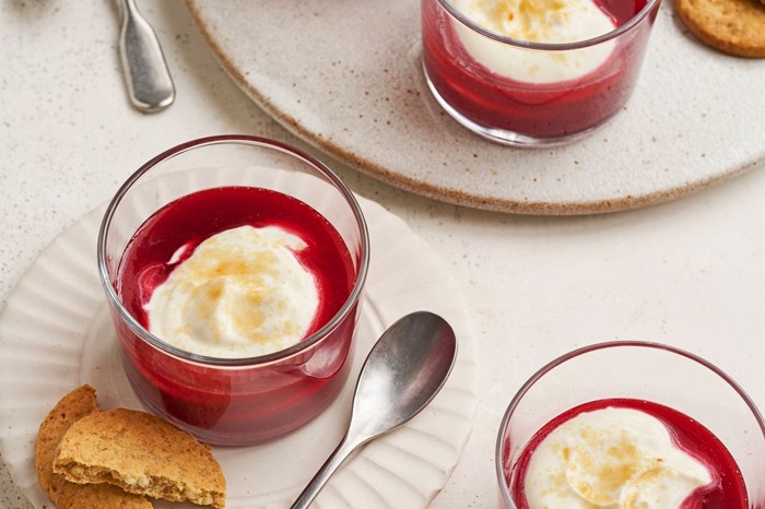 Four glasses filled with berry coulis and topped with yogurt and double cream on plates next to halved ginger biscuits.