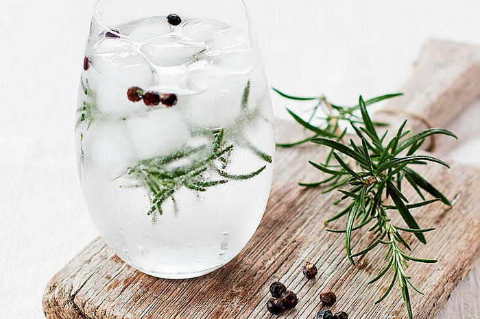 a glass of gin and tonic with ice, with juniper berries floating in the liquid and a wooden chopping board underneath