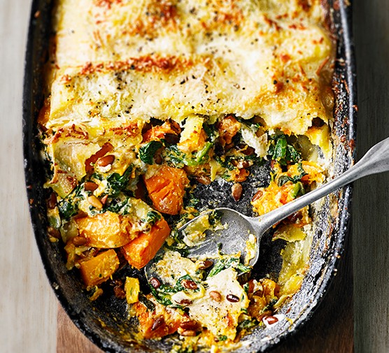 Butternut squash, spinach and mascarpone lasagne in a large oval black baking dish, placed on a wooden plank