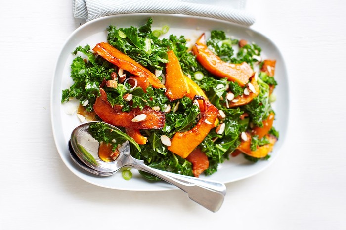 Kale salad with roasted butternut squash, pomegranate molasses and almonds on a white plate with serving spoons