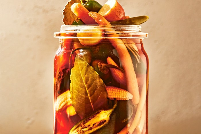 a glass jar on a stone surface overflowing with chillies, bay leaves, sliced potatoes and baby corn