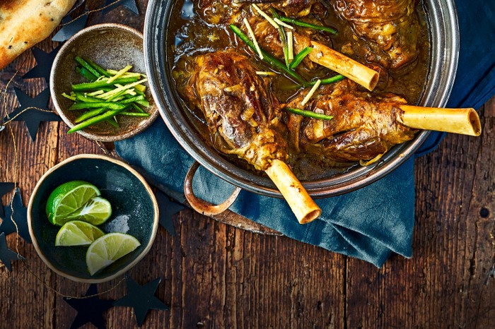 A dish filled with lamb shanks in a deep brown curry broth with lime wedges on the side