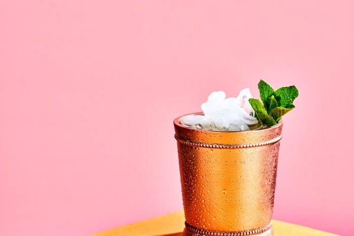 A copper cup filled with crushed ice and a sprig of mint on a yellow table with a pink background