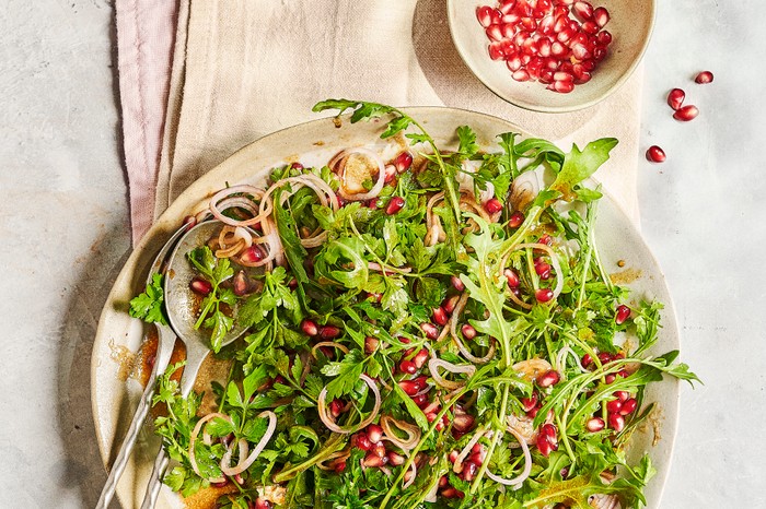 A round white serving plate of green leaves and red pomegranate seeds, sat on a folded piece of white linen