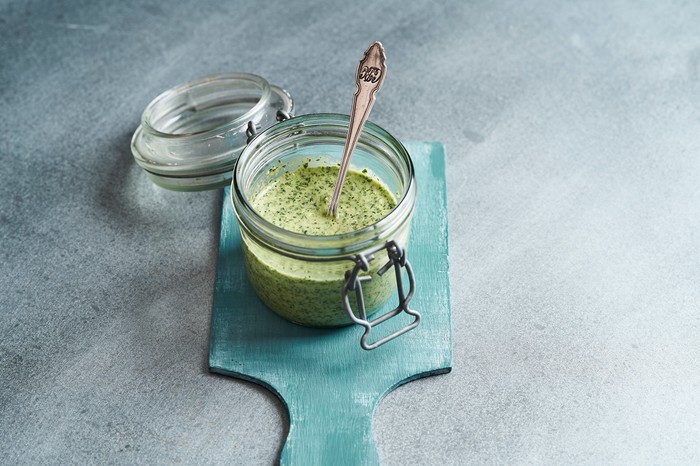 A grey background with a blue board, topped with a jar of green pesto sauce