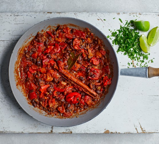 a silver pan with a wooden handle on a white wooden chopping board filled with pan of a bright-red coloured curry