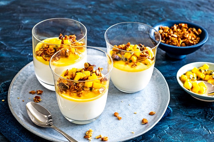 A grey plate holding three glasses filled with white mousse and topped with mango and granola, on a dark blue background