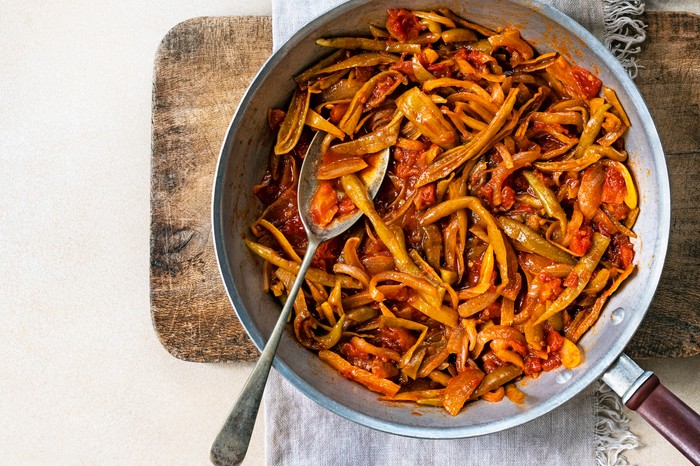 A steel frying pan with a spoon filled with orange and red slow cooked peppers and tomatoes