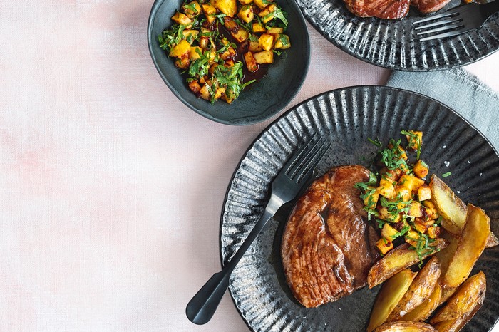 Two dark grey plates topped with gammon steaks, potato wedges and chunks of pineapple with green herbs