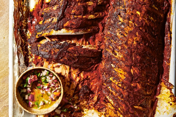 two racks of pork ribs on baking parchment in a roasting tray with a pot of salsa