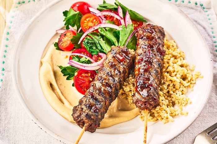 a plate of hummus, salad and lamb kebabs on wooden skewers