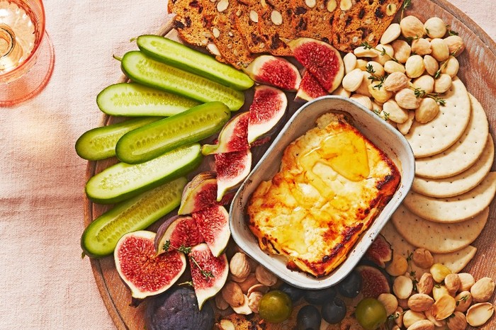 A baked block of feta on a board with figs, crackers, almonds and sliced cucumbers