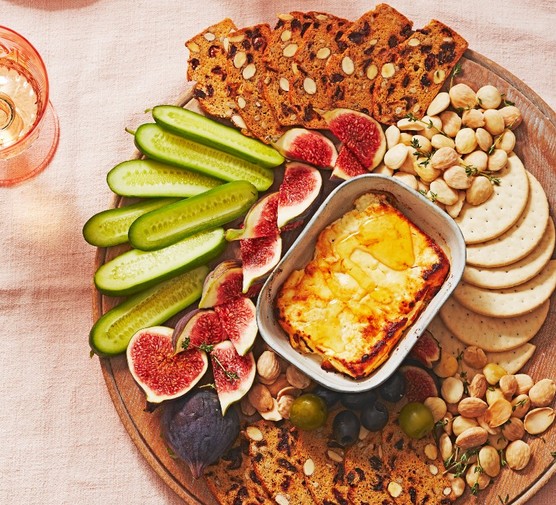 A baked block of feta on a board with figs, crackers, almonds and sliced cucumbers