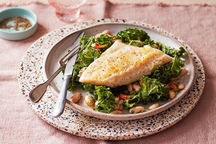 A plate of cod and kale on a terrazzo board with a pink background and glass of water