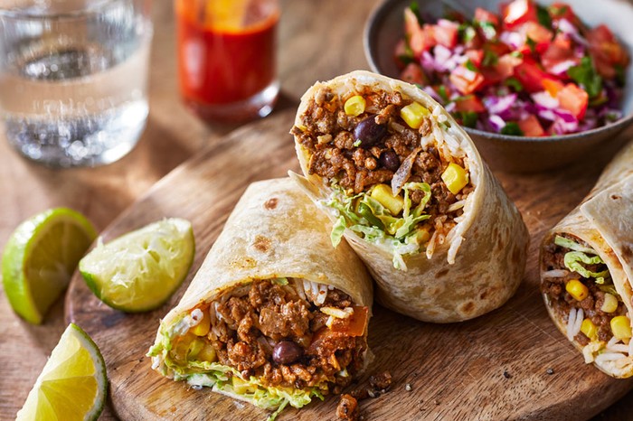 Halved burritos on a wooden board next to limes and a bowl of salsa