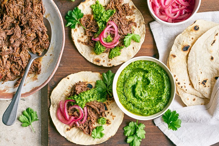 Two tacos filled with beef cheek, salsa, pickled onion and topped with coriander. Next to a pile of tortillas and a bowl of beef cheek.