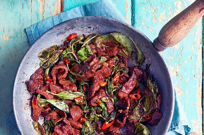 A beef stir-fry in a metal wok with a wooden handle on a blue background