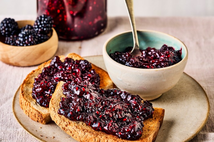 A glass jar of deep purple jam with a pot and a spoon, with two slices of toast covered in jam on a ceramic plate