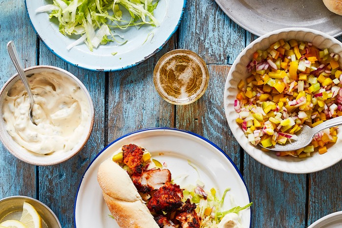 Various plates scattered over a washed blue wooden surface, filled with a sub roll of blackened cod, lettuce, salsa and creamy sauce