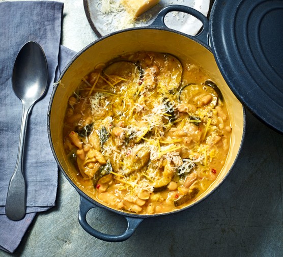 Braised Courgette & Cannellni Beans V2 A cast iron pot of creamy braised courgette with the lid leaning on the side, on a grey concrete background with a grey napkin and brushed sliver serving spoon