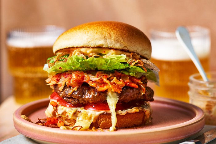 A burger layered with kinchi and salad sat on a pink plate with a glass of lager in the background