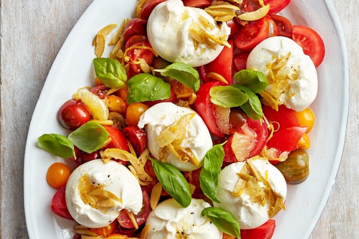 A white oval platter filled with burrata, olives and tomato salad on a whitewashed wooden background