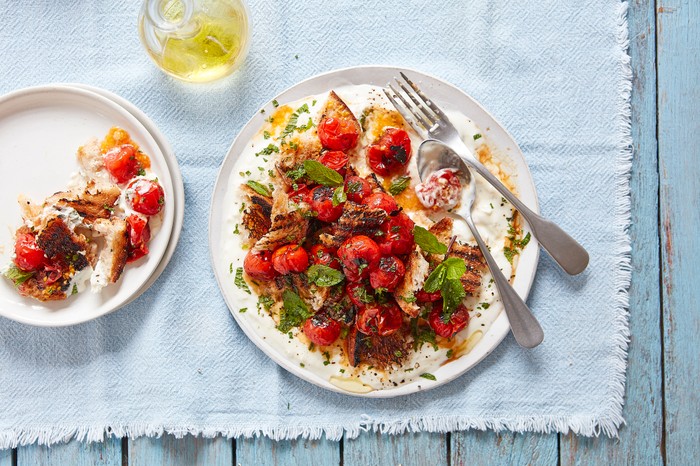 A white plate of charred tomatoes piled on a bed of yogurt with a spoon and fork, sat on a pale blue cloth