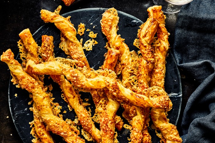 Golden baked cheese straws on a black plate with a black linen cloth and a glass of white wine