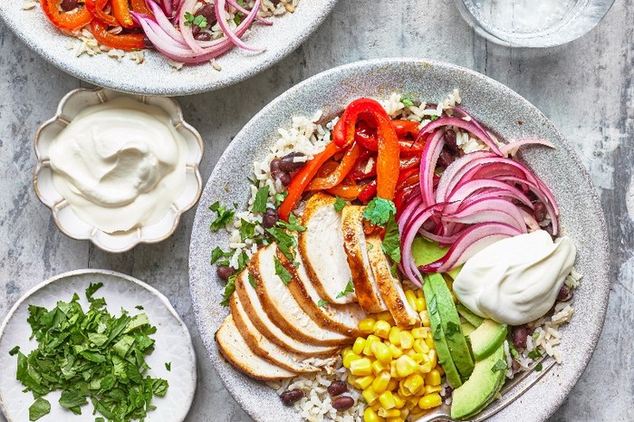 A bowl filled with sliced chicken, veg and rice next to a small plate of chopped herbs