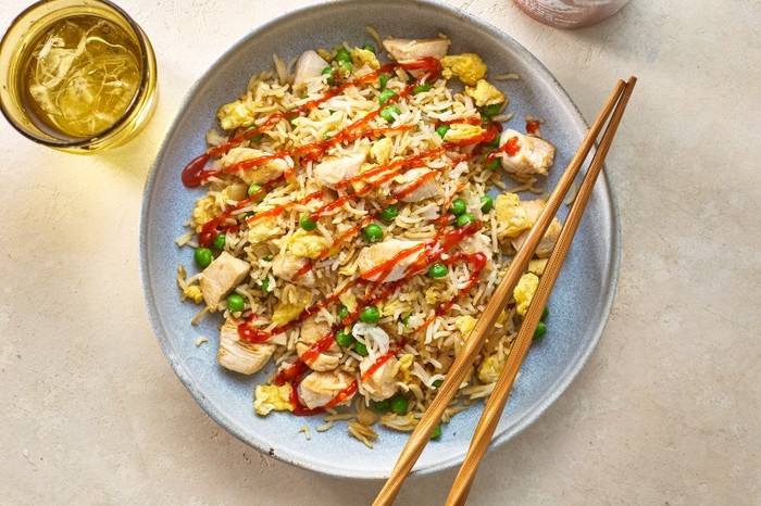 a bowl of fried rice with chunks of chicken and peas topped with hot sauce and a pair of chopsticks