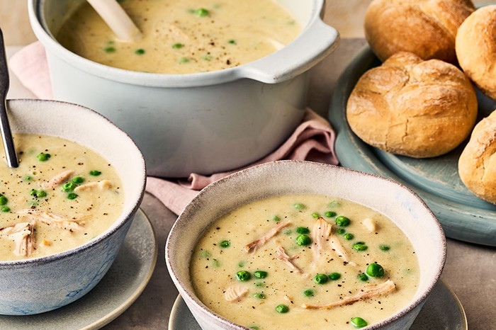 A pot and two bowls of chicken soup next to a board of bread rolls