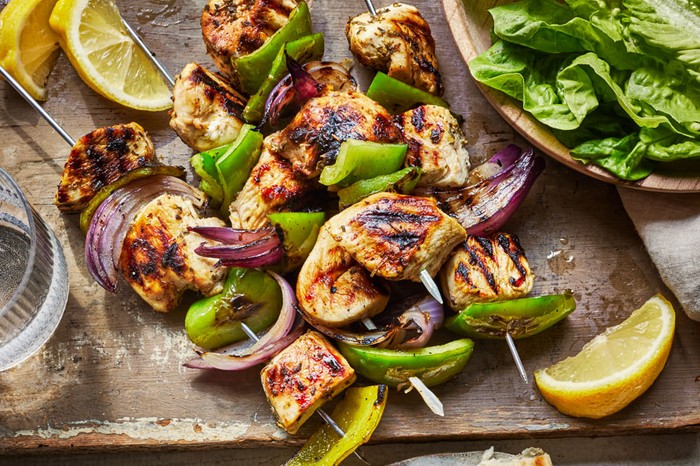 Chicken souvlaki skewers on a board, with a bowl of salad leaves on the side and two torn grilled pitta breads