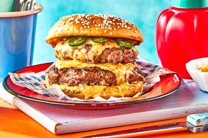 A double patty burger in a seeded bun with melted cheese on a red plate in front of a ketchup bottle