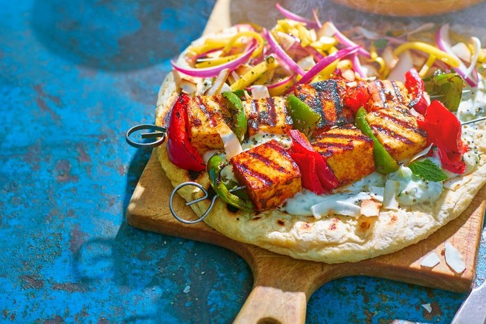 A round flatbread topped with bbq paneer skewers on a wooden board with a blue background