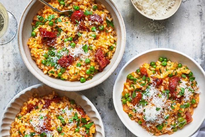 Three bowls of chorizo and pea risotto on a grey table with a pot of grated parmesan