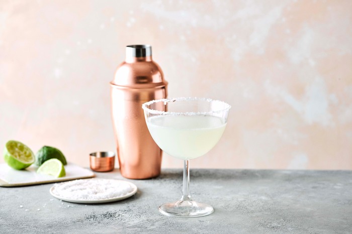 Margarita in a glass with copper cocktail shaker and a plate of limes behind