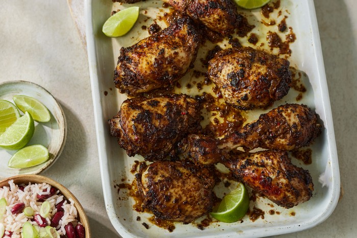 Jerk chicken with wedges of lime and in a white roasting tin next to a small bowl of rice