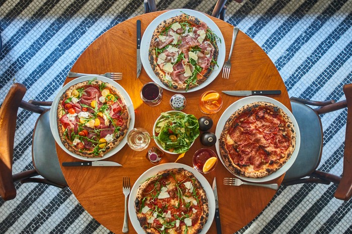 A mahogany table is topped with four pizzas and glasses of Aperol spritz at Soho House's Cecconi's Pizza Bar