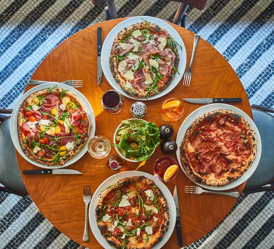 A mahogany table is topped with four pizzas and glasses of Aperol spritz at Soho House's Cecconi's Pizza Bar