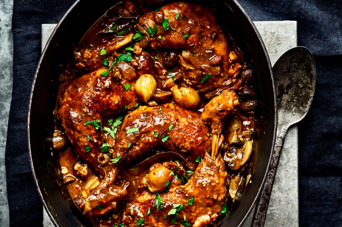 A black oval casserole pot with a serving spoon, filled with coq au vin and topped with parsley