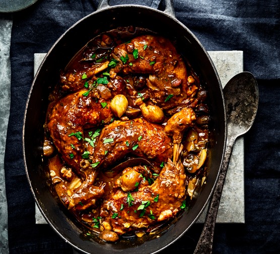 A black oval casserole pot with a serving spoon, filled with coq au vin and topped with parsley