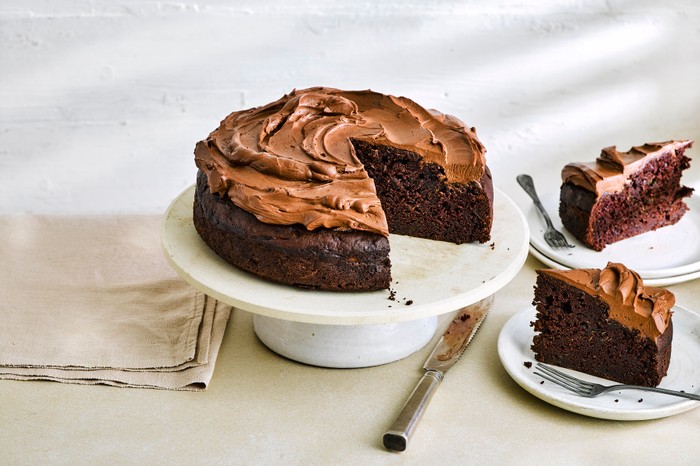 chocolate cake topped with chocolate frosting on a ceramic cake stand with a slice cut out of the middle