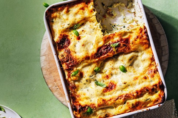 A white ceramic oven dish filled with baked lasagne on a wooden board and a green surface