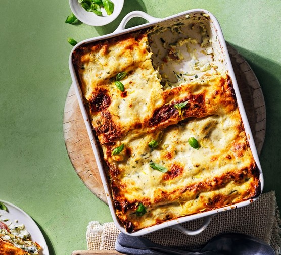 A white ceramic oven dish filled with baked lasagne on a wooden board and a green surface