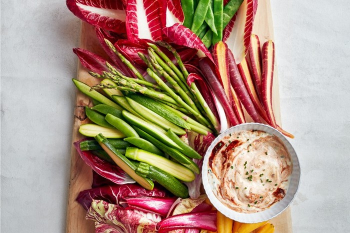 A wooden board on a grey stone background flled with vegetable crudités and a small pot of chipotle aïoli with red rippled through